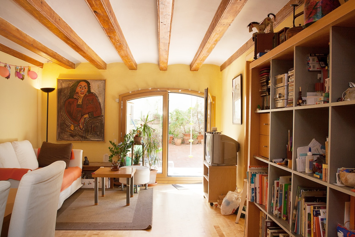 A welcoming living area features a comfortable white sofa paired with an orange accent cushion. A small coffee table sits in front, surrounded by plants and bookshelves. Natural light floods in through double doors leading to a private terrace, enhancing the space's inviting feel.