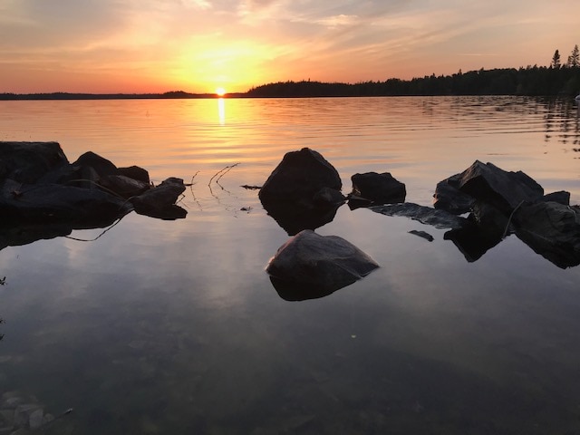 A tranquil sunset view over a calm lake is depicted, with the sun positioned just above the horizon. Smooth stones along the water's edge are visible, reflecting the warm colors of the sky and creating a serene atmosphere.