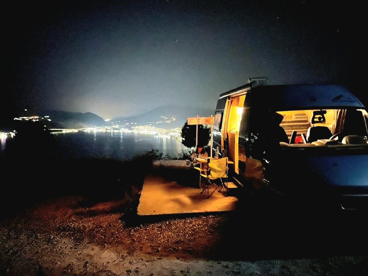 A camping van is positioned on a hillside, illuminated from within. Two folding chairs are set outside, facing a serene view of twinkling city lights reflecting on the water at night. Mountains are faintly visible in the background under a starry sky.