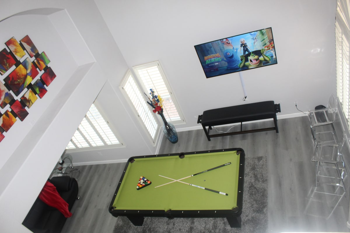An overhead view of a game room features a green pool table with positioned cues and balls. A large smart TV is mounted on the wall, displaying animated content. Decorative artwork adorns the wall, complemented by a small seating area with modern furnishings.