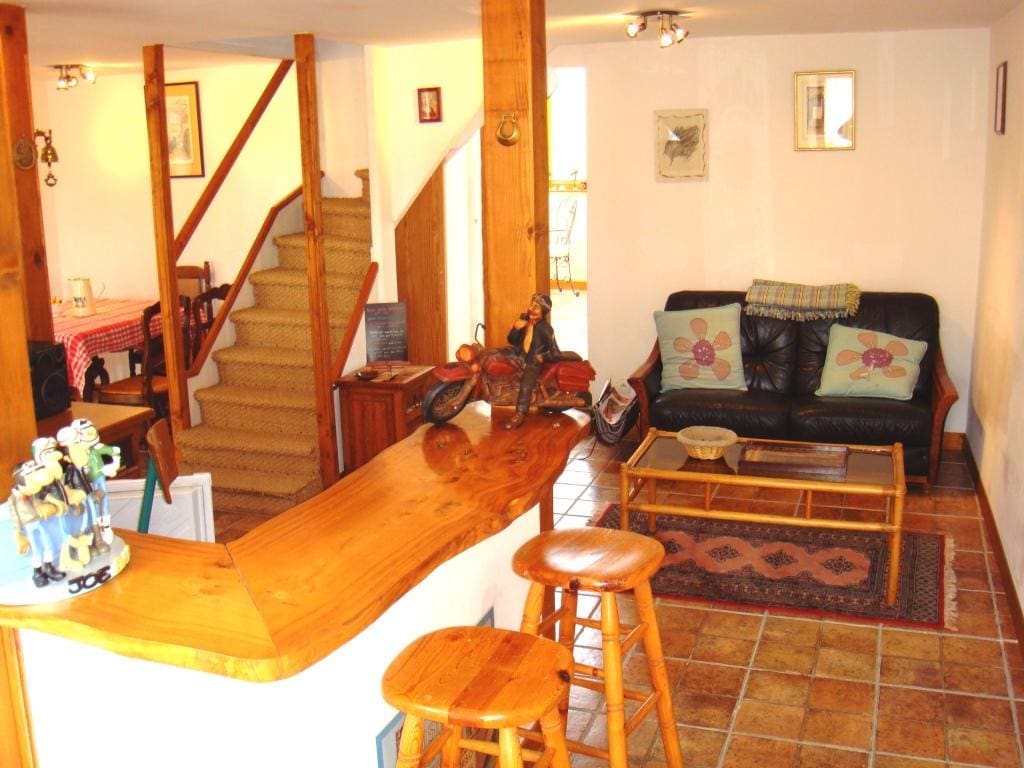 An open living area is featured, showcasing a wooden bar with high stools and a coffee table. A black leather couch adorned with floral cushions is present, along with warm lighting and decorative art on the walls. A staircase leads to the upper level.