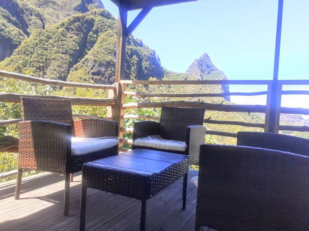 A seating area is featured on the terrace, equipped with four woven chairs and a low table. The backdrop displays a panoramic view of towering mountains and lush greenery, enhancing the outdoor space's inviting atmosphere.