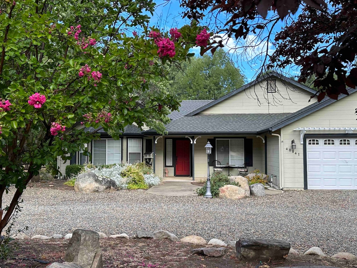 The exterior of a charming house is framed by blooming flower trees and landscaped shrubs. A gravel driveway leads to a welcoming entrance, featuring a red front door and large windows. Smooth stones are arranged along the front, providing a natural border.