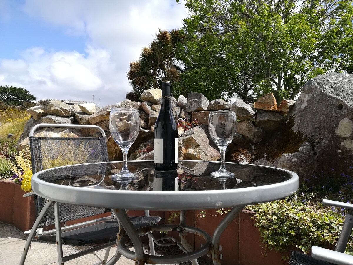 A round glass table is set with two wine glasses and a bottle of wine, placed against a backdrop of natural stone landscaping and greenery. The scene is illuminated by soft daylight, highlighting the tranquility of the outdoor space.