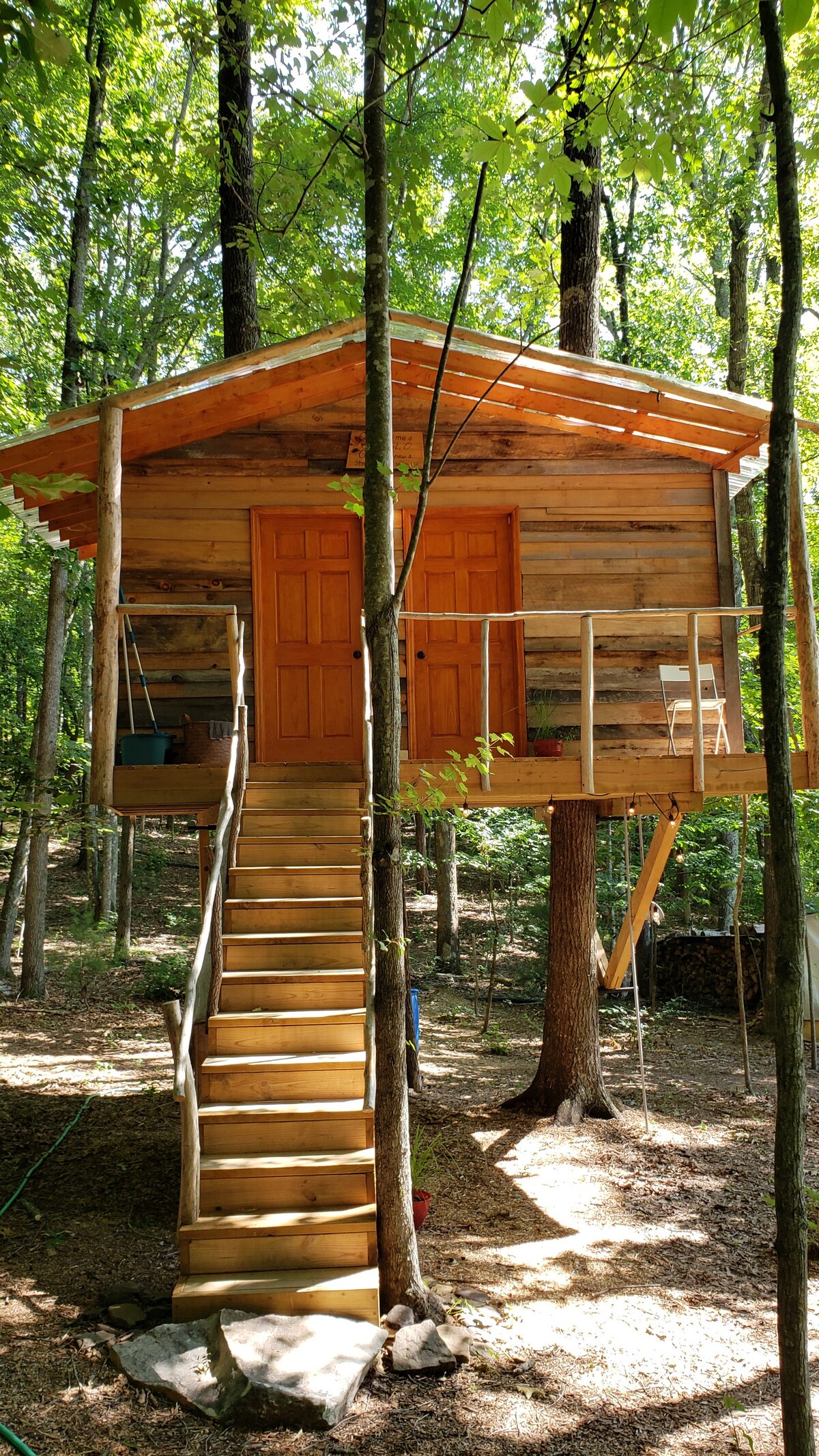 A rustic treehouse is elevated on wooden stilts, accessible by a set of steps. The exterior features natural wood siding, with a pair of orange doors centered at the front. Surrounding greenery provides shade, highlighting the treehouse’s secluded setting.