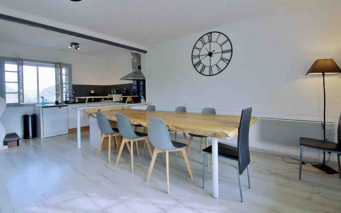 A spacious dining area features a large, natural wood table surrounded by several grey and black chairs. A large clock is mounted on the wall, while a modern lamp stands nearby. Natural light enters through multiple windows, enhancing the inviting atmosphere.