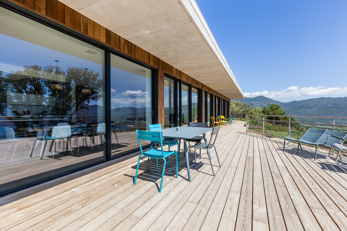 The spacious terrace features wooden flooring and is furnished with colorful chairs and a dining table. Floor-to-ceiling windows provide abundant natural light, while the surrounding landscape showcases rolling hills and distant mountains under a clear blue sky.