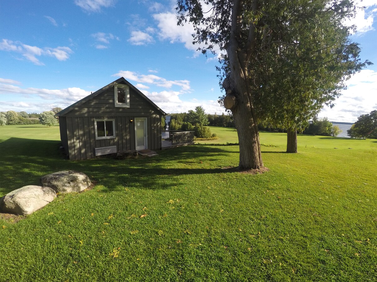 A charming cottage is set against a vast green lawn, with large rocks and a tree in the foreground. Light clouds drift across a blue sky above, while the serene landscape extends towards the horizon, hinting at the nearby lake.