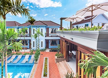 A landscaped outdoor area features a swimming pool surrounded by lounge chairs. Nearby, a stylish shaded terrace is visible, providing a space for relaxation and dining. Lush tropical plants decorate the space, contributing to a vibrant atmosphere under a clear sky.