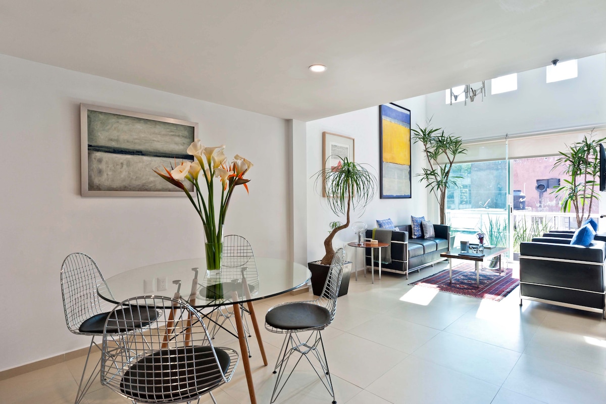 An open-plan living and dining area is illuminated by natural light from large windows. A glass dining table with modern chairs is placed next to a cozy seating area, featuring two black armchairs and decorative plants. Artwork adorns the walls, contributing to the contemporary ambiance.