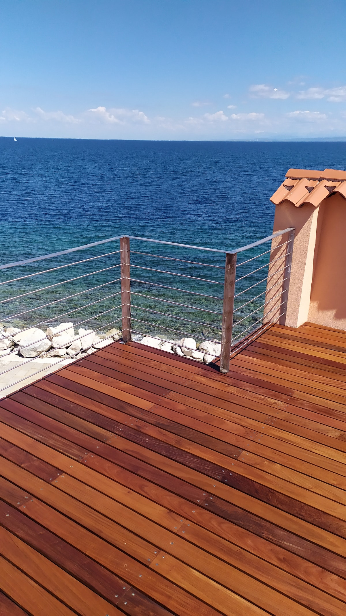 A wooden terrace extends towards the sea, featuring smooth planks and a secure railing. The tranquil waters are visible nearby, displaying shades of blue and green, with rocks lining the shoreline. A bright sky above enhances the area's natural beauty.