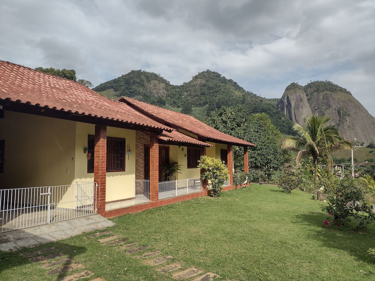 A peaceful outdoor view of a single-story accommodation, featuring a brick facade and a red-tiled roof. The surrounding landscape includes lush greenery and distant mountains, framed by a neat lawn with a pathway leading to the entrance.