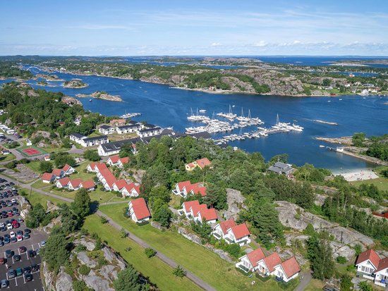 An aerial view captures a scenic coastal landscape featuring a marina filled with boats. Nearby, clusters of white cottages with red roofs are surrounded by lush greenery. The shoreline is visible with a sandy beach area, and parking spaces are noted in the foreground.