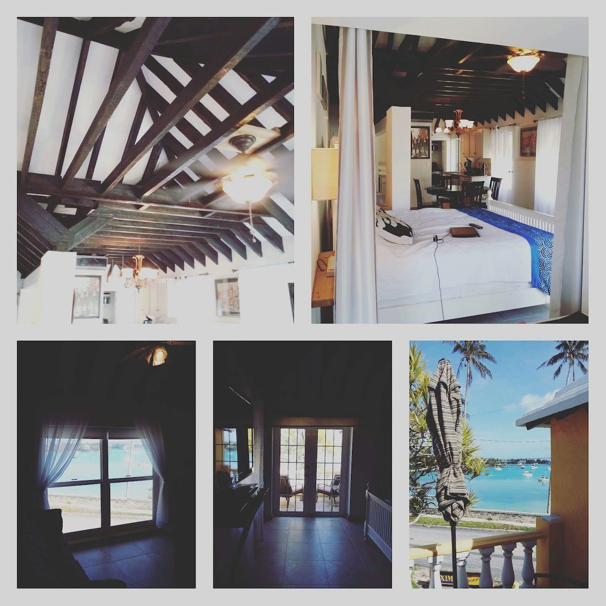 The interior space features wooden beams overhead and a ceiling fan. A spacious bed is visible, offering comfort and relaxation. Large windows allow natural light, showcasing a glimpse of the ocean view outside. Additional seating and a dining area are present, emphasizing functionality.