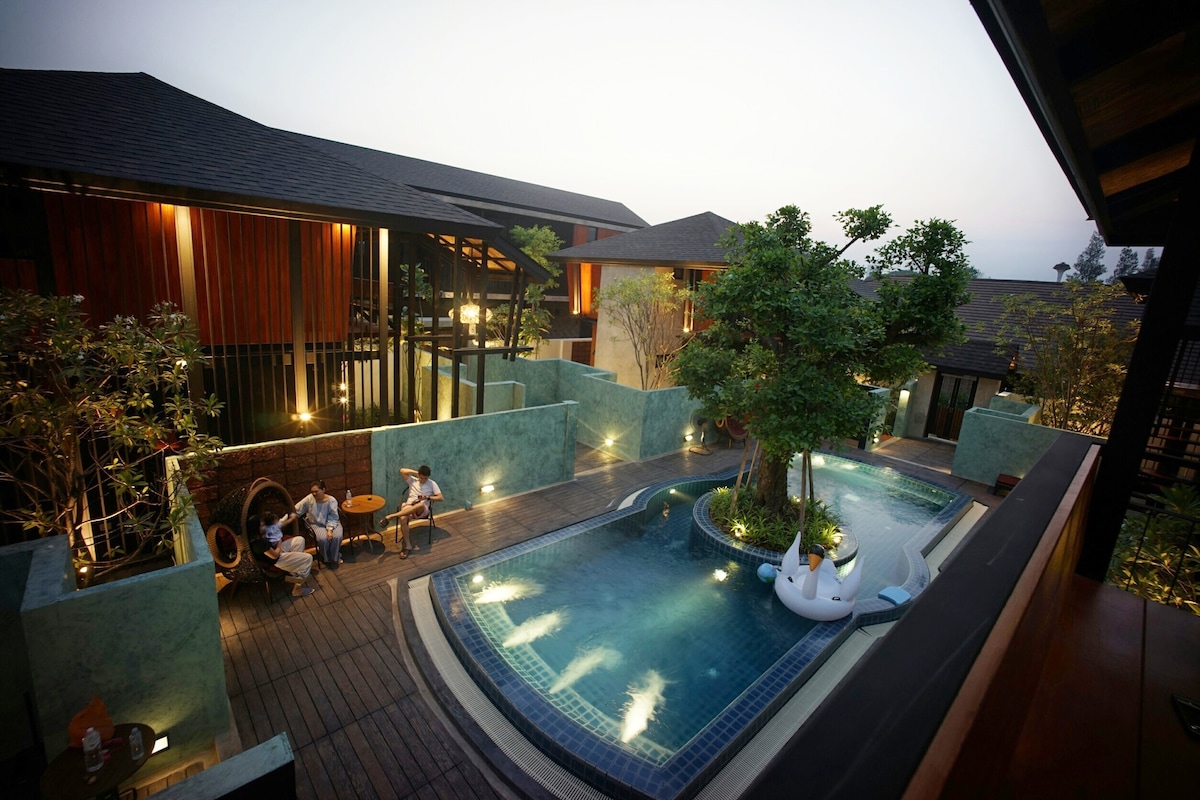An open-air swimming pool is surrounded by a wooden deck and vibrant greenery. Several loungers are positioned by the water, offering relaxation options. Nearby, guests are seated at a small table, enjoying the serene environment as the evening sky transitions to twilight.