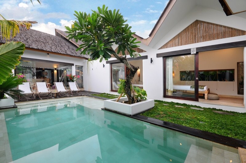 An inviting outdoor area features a clear blue pool surrounded by green grass and tropical plants. Several lounge chairs are arranged neatly beside the pool, with well-maintained trees providing a sense of tranquility. The surrounding architecture showcases a blend of traditional and modern design.