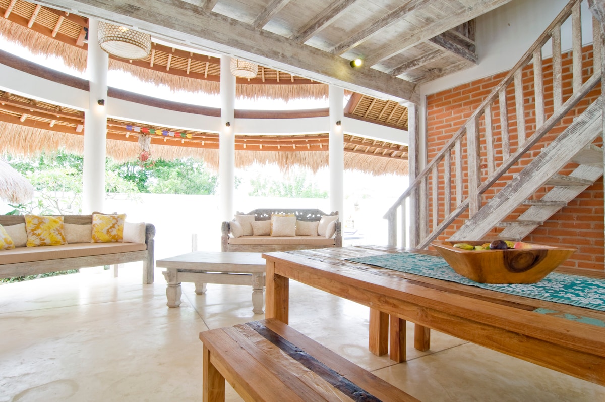 An open living area is featured with a wooden dining table at the center, complemented by a bowl of fresh fruit. Comfortable seating is arranged nearby, with colorful pillows on a light sofa. Natural light floods through round windows and a thatched roof enhances the tropical feel.
