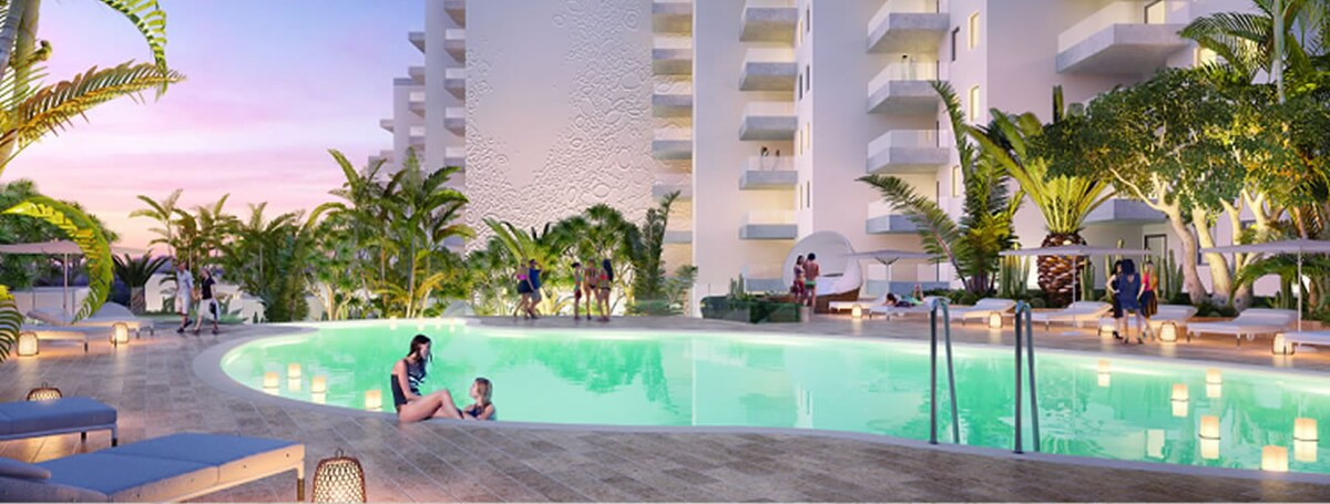 A heated pool is surrounded by lush tropical landscaping and modern architecture. Sun loungers are positioned along the poolside, while people can be seen enjoying leisure time in and around the water. Soft lighting enhances the serene atmosphere as the sun sets in the background.