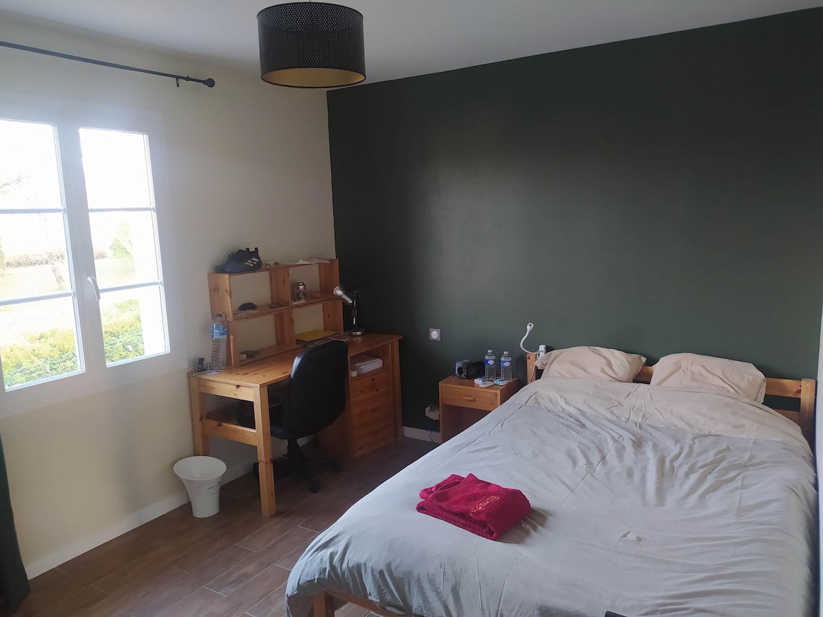 A spacious bedroom features a wooden bed with neatly arranged linens and a red towel. A black desk with a chair is positioned against the wall, accompanied by a small wooden nightstand and a wastebasket. Natural light enters through a window with a simple treatment.
