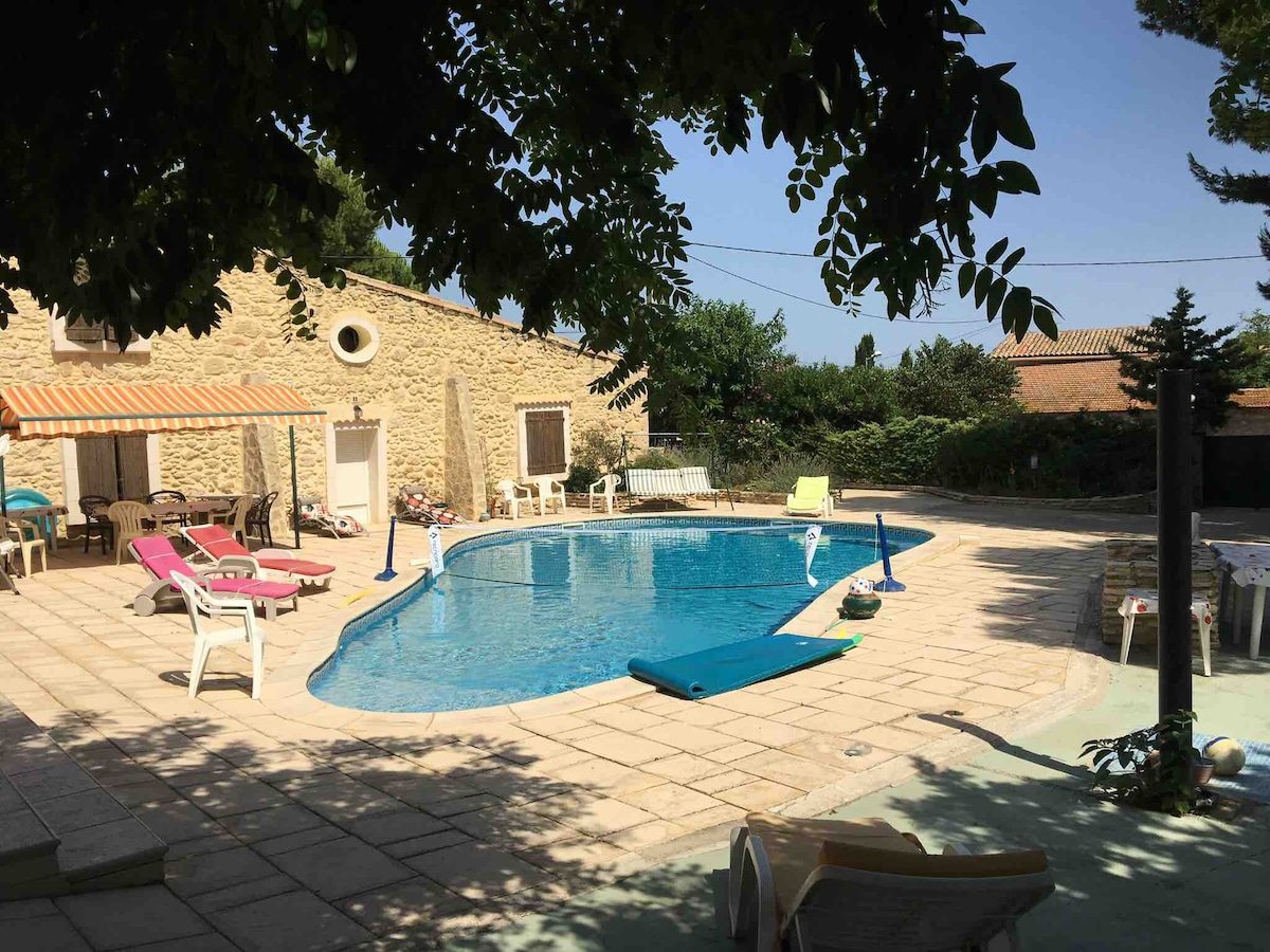 A tranquil outdoor area features a spacious swimming pool surrounded by sun loungers and shaded seating. The villa is visible in the background, complemented by greenery. Umbrellas and poolside decor add comfort. A clear blue sky enhances the serene atmosphere.