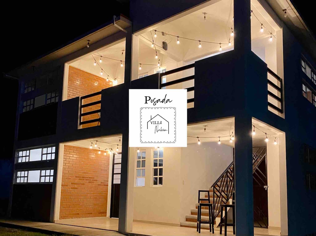 The exterior of the two-story villa is illuminated by warm string lights, creating a welcoming ambiance. Large windows showcase the indoor areas, while a staircase leads to the upper level. The contemporary design features a mix of brick and white walls.