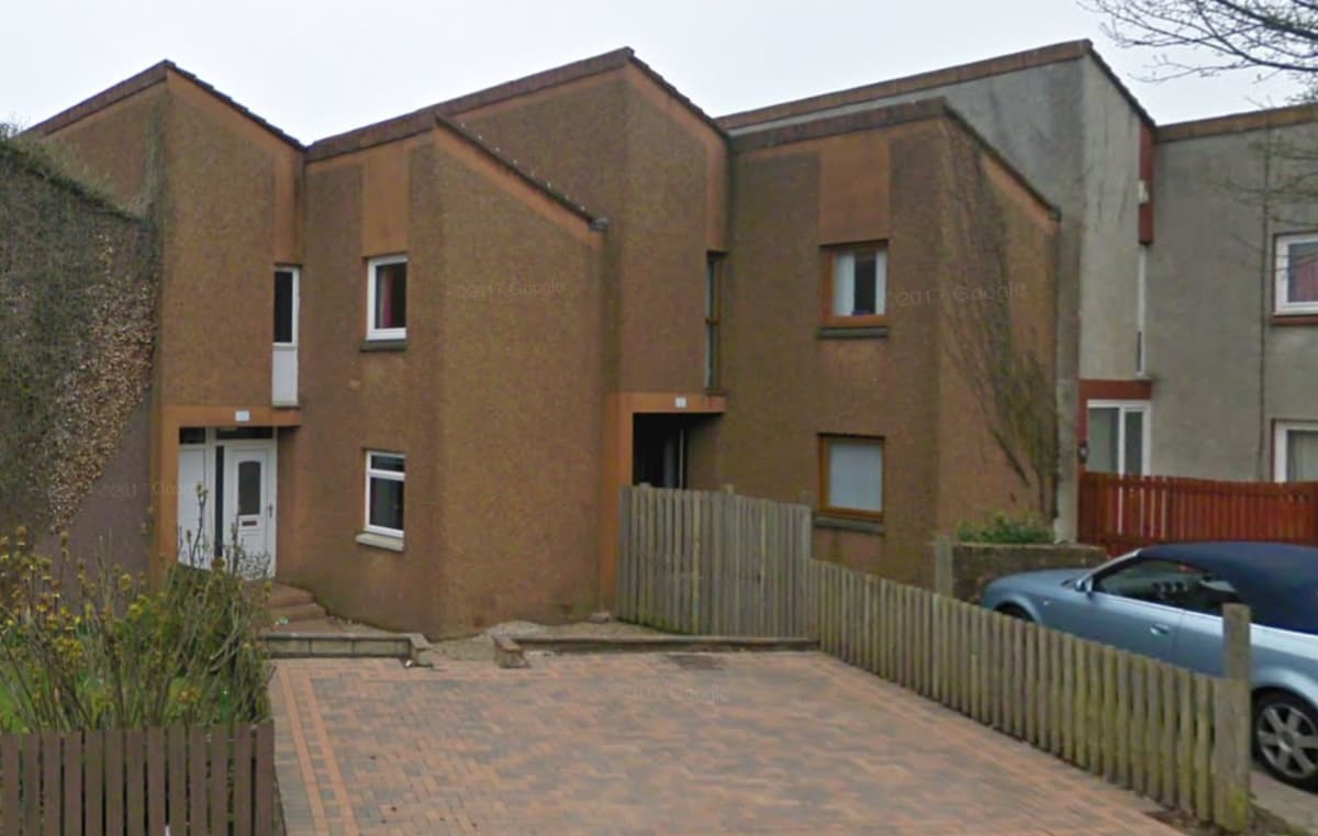 The image shows a modern home with an angular design, featuring a two-tone exterior of textured brown and beige. A paved drive leads to the entrance, while a fence borders the front garden. A parked car is visible beside the property, contributing to the suburban setting.