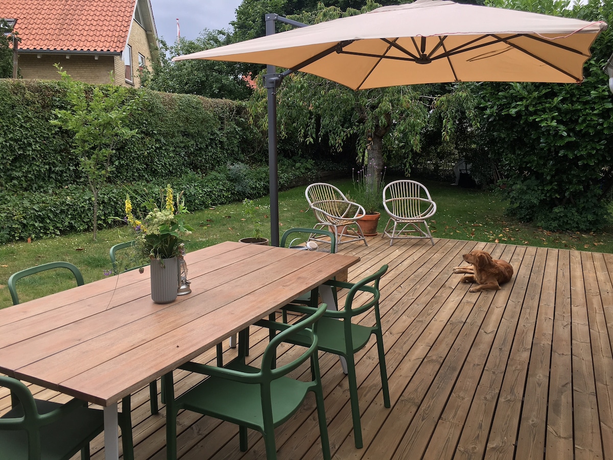 A spacious wooden deck features a large dining table surrounded by green chairs, under a shaded umbrella. In the background, a well-maintained garden is visible, with various plants and trees. A relaxed dog can be seen resting on the deck.