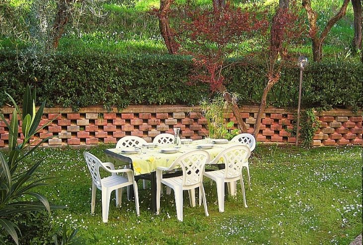An outdoor seating area is set within a shared garden, featuring a table with a yellow tablecloth surrounded by white plastic chairs. Lush greenery and flowering plants create a serene atmosphere, while a low brick wall provides structure at the back.