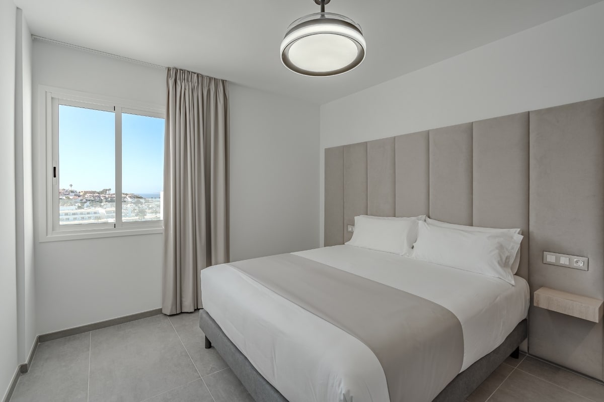 A modern bedroom features a spacious bed with a neutral-colored headboard. Soft, natural light enters through a large window, accompanied by light curtains. Neutral tones and simple decor contribute to a serene environment, offering views of the surrounding area beyond the glass.