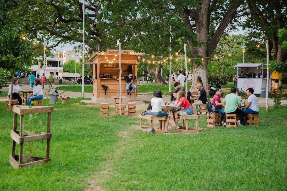 An outdoor space is captured, showcasing a vibrant gathering area with people seated at rustic wooden tables. String lights create a welcoming ambiance above a small bar kiosk. Lush green grass surrounds the setting, complemented by large trees providing shade.