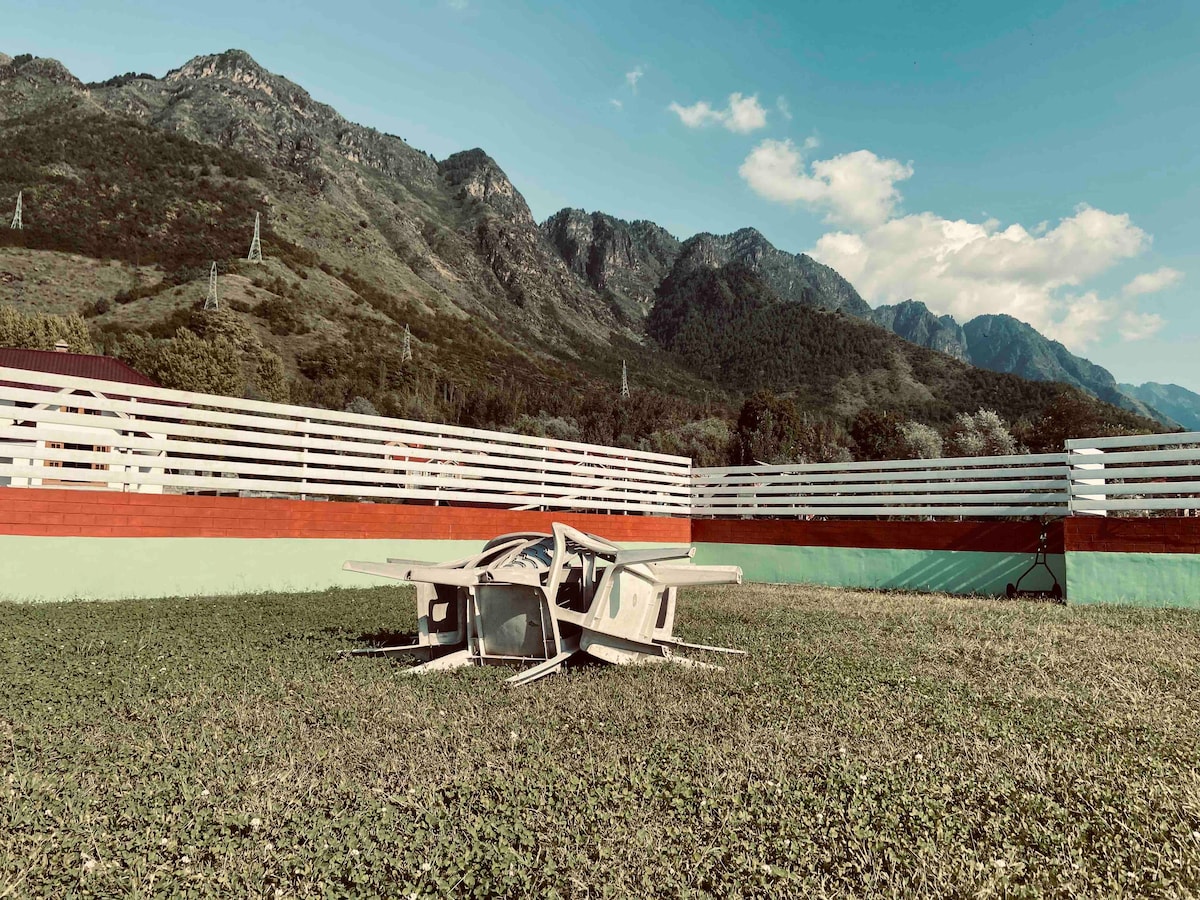 An outdoor area featuring a grassy lawn is framed by a low white fence. In the foreground, a unique wooden structure resembling a small shelter is positioned on the grass. The backdrop reveals the stunning Zabarwan mountains under a clear blue sky.