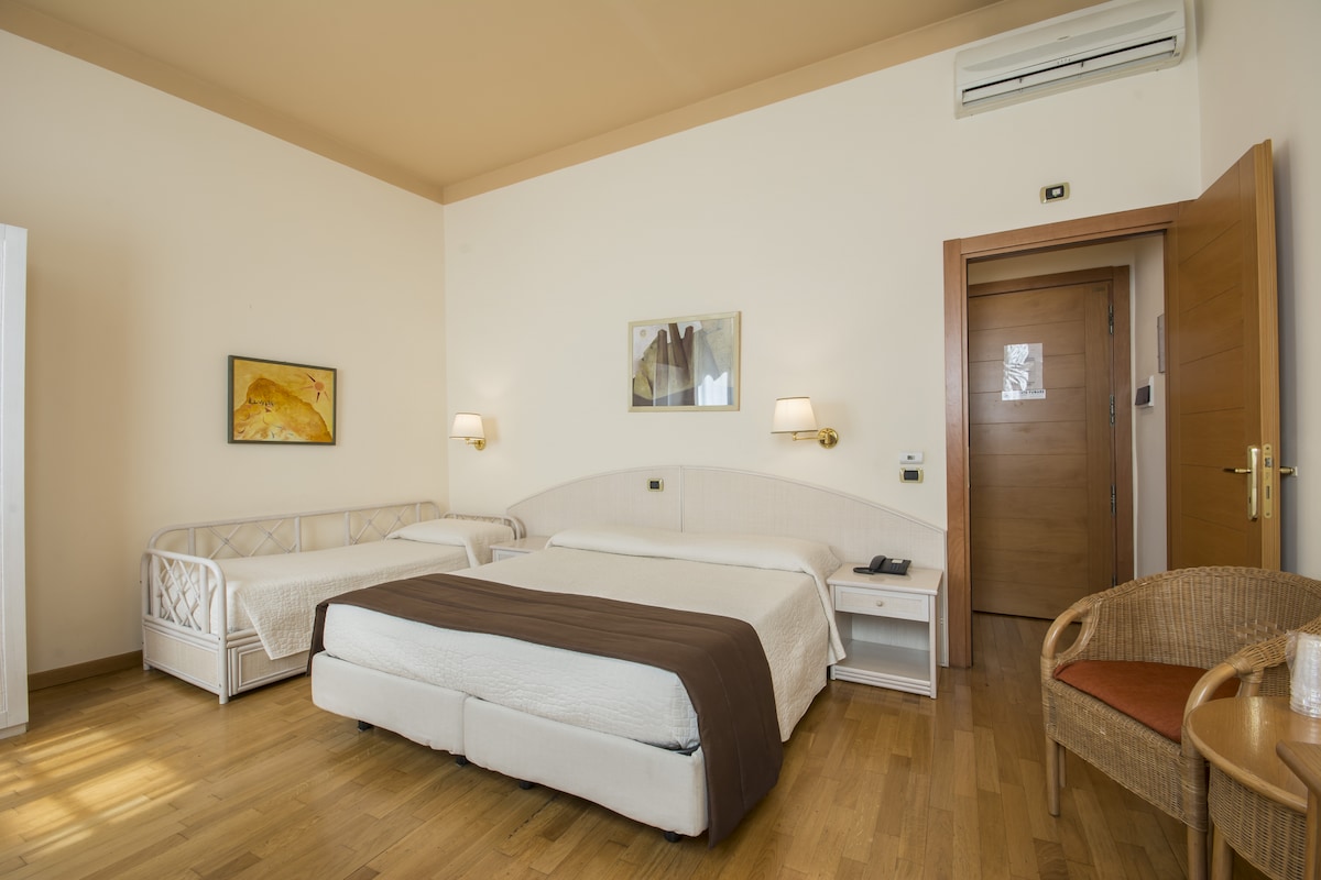 A spacious room features a double bed and a single daybed, complemented by modern décor. Light-colored walls and wooden flooring enhance the bright atmosphere. Two bedside lamps provide soft lighting, while a seating area includes a woven chair. A door leads to the corridor.