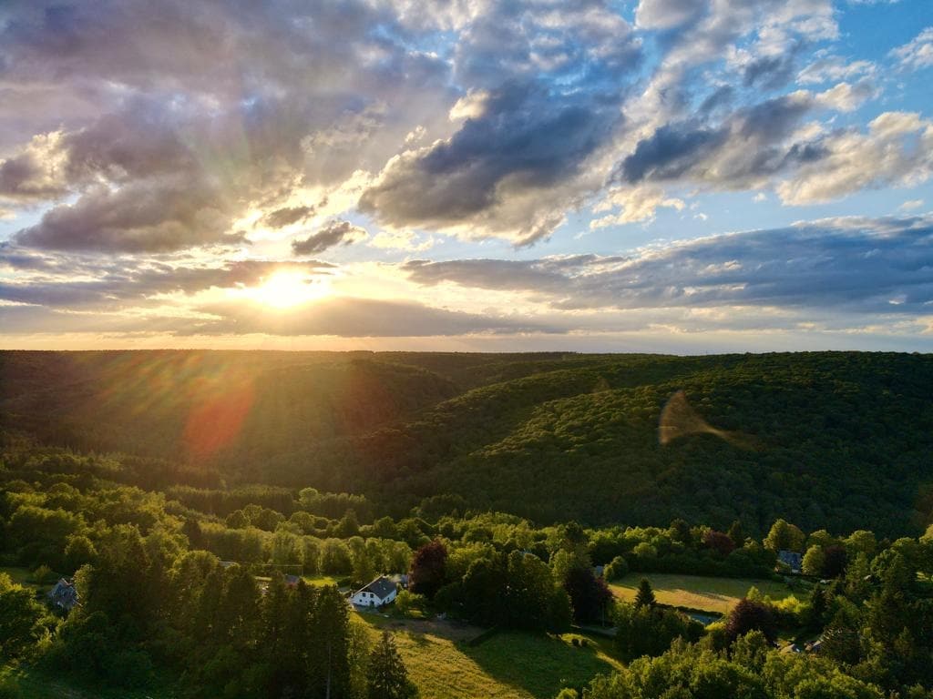 Aerial view captures rolling hills blanketed with lush greenery under a vibrant sky. The sun is seen setting on the horizon, casting warm hues across the landscape. A charming white house nestled among trees is visible in the foreground, highlighting the area's natural beauty.