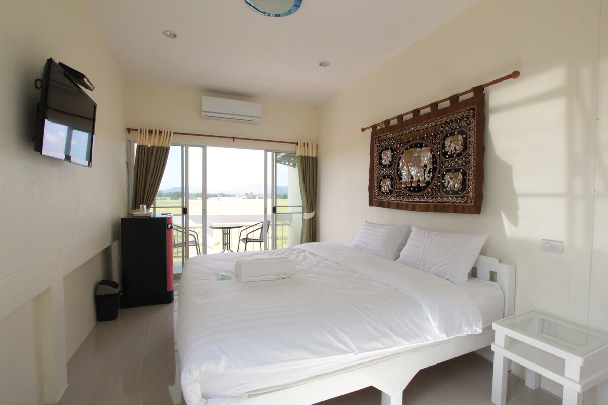 A spacious studio room features a large bed with crisp white linens and a decorative wall hanging. A flat-screen TV is mounted on one wall, while a glass door opens to a balcony offering views of the surrounding landscape. A small fridge and wastebasket are visible.