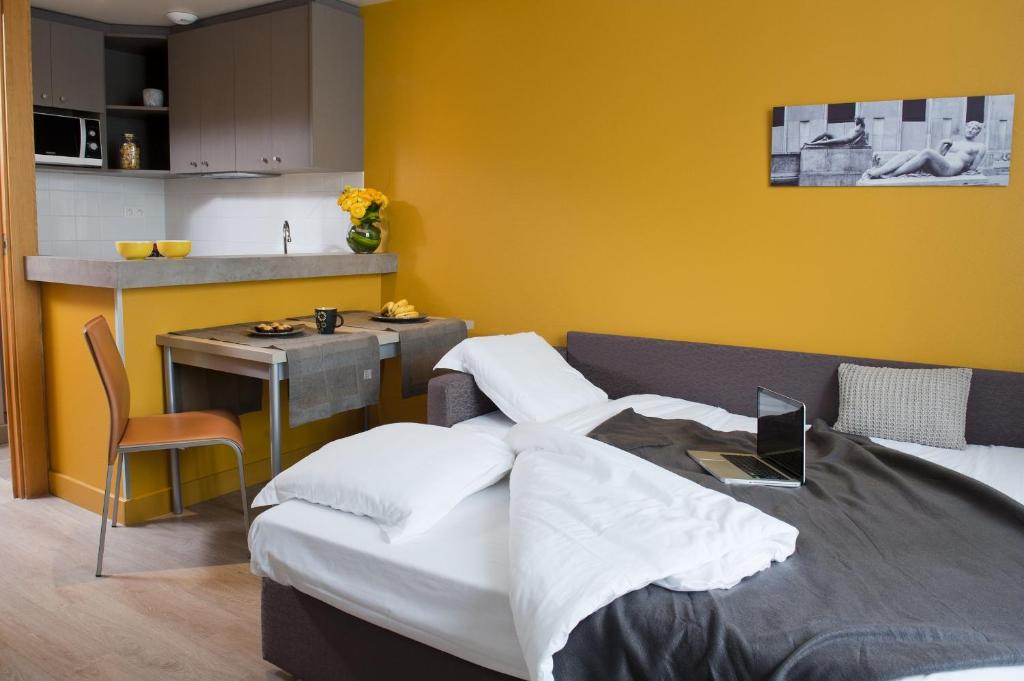 A studio apartment features a spacious bed with white linens and a grey throw. A small table with a grey tablecloth is set for breakfast, with a laptop on the bed. The kitchen area is equipped with modern appliances, and bright yellow walls add warmth to the space.