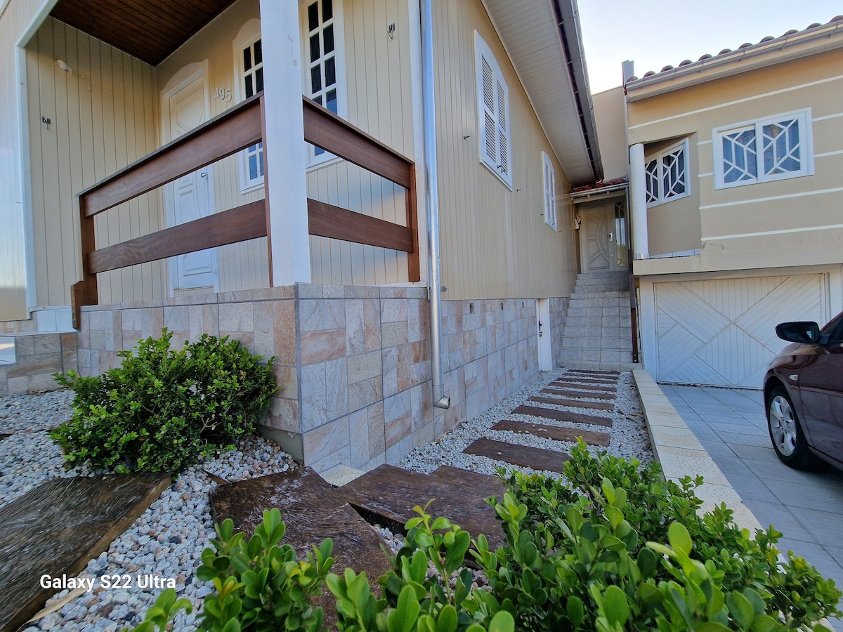 The entrance of the home is framed by a wooden railing and features a stone pathway leading to the stairway. Lush greenery and gravel accentuate the surrounding area, while a garage door is visible in the background, complementing the welcoming exterior.
