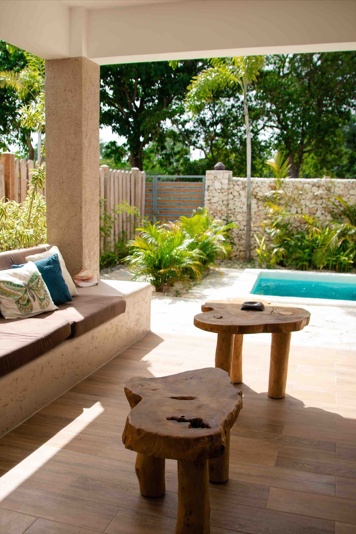 A shaded outdoor seating area features a comfortable bench with soft cushions and two wooden side tables. Sunlight filters through foliage, illuminating the space. A small pool can be seen nearby, surrounded by tropical plants and a stone wall.