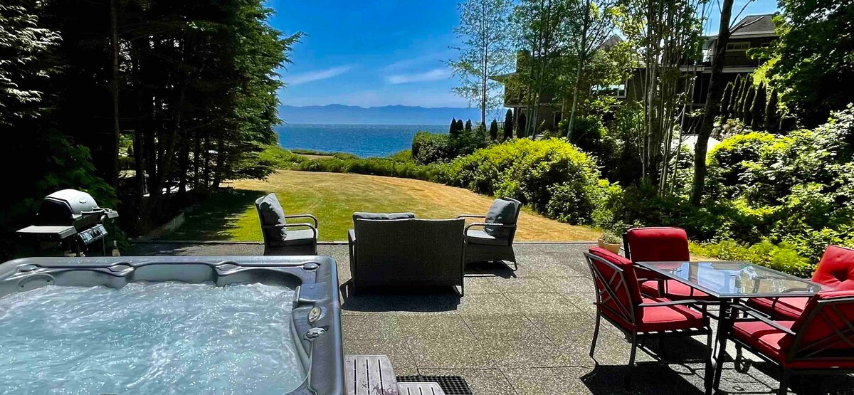 An outdoor space features a hot tub and patio furniture set, including a dining table with chairs. Lush greenery surrounds the area, leading to a clear view of the ocean and distant mountains under a bright blue sky.