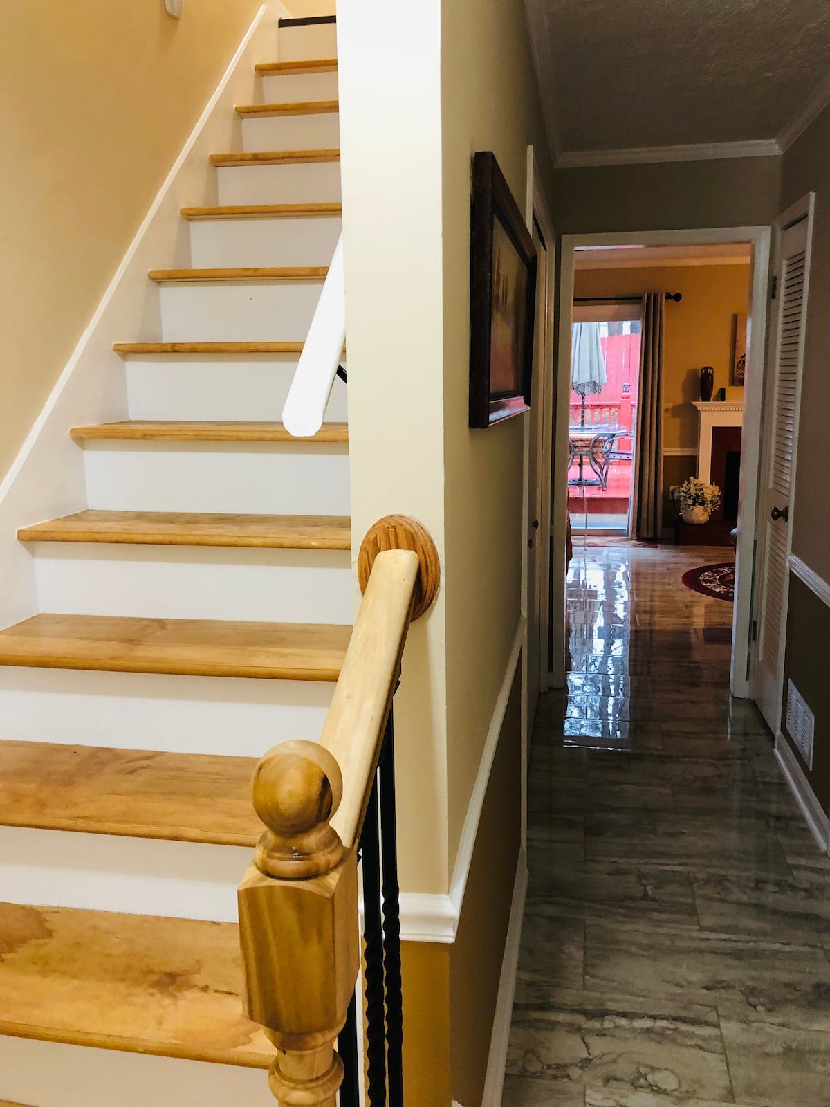 A staircase with light wooden steps leads to the upper level, complemented by a wooden handrail. A hallway is visible in the background, featuring tiled flooring and doorways to adjacent spaces. Soft natural light enters from the nearby windows.