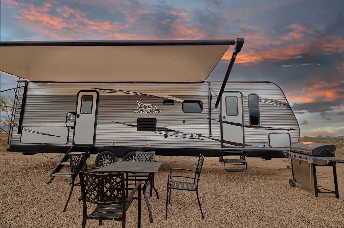 A spacious camper is positioned under a large awning, providing shade over outdoor seating. A barbecue grill is visible nearby. The landscape features dramatic sunset hues, with distant mountains creating a serene backdrop.