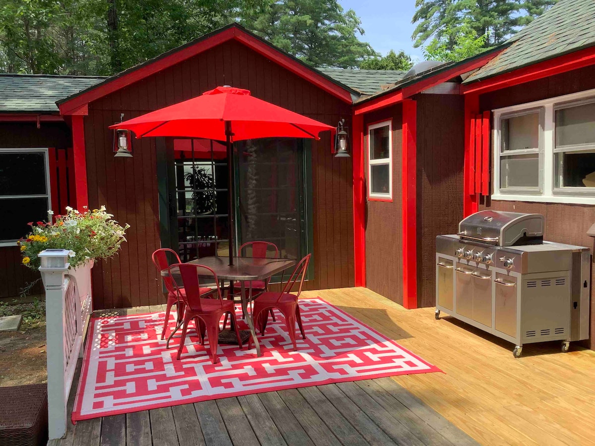 An outdoor dining area is presented with a round table surrounded by four red metal chairs, situated on a patterned rug. A bright red umbrella provides shade, and a gas grill is positioned nearby, enhancing the inviting atmosphere for outdoor meals.