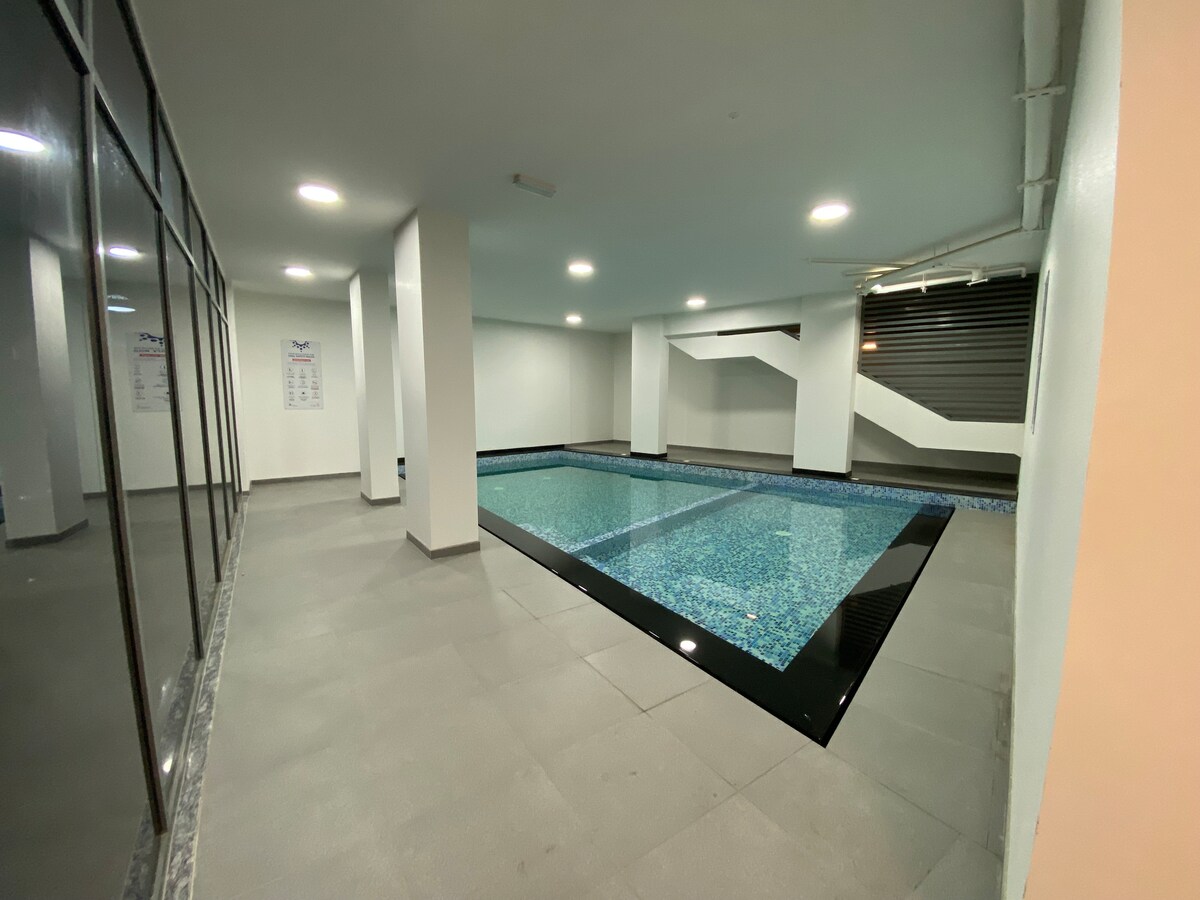 A spacious indoor pool area features a rectangular swimming pool with a blue mosaic tile trim and a dark edge. White walls and bright ceiling lights enhance the open atmosphere. Large windows provide a view of the nearby room and natural light.