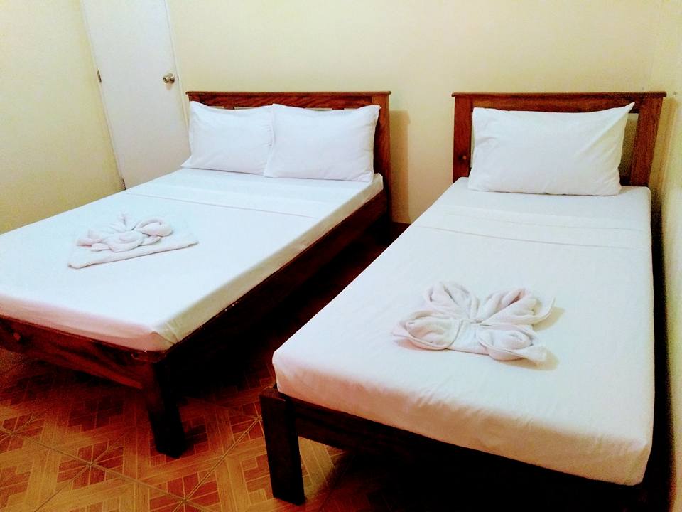 The room contains two wooden beds, one double-sized and one single-sized, both dressed in crisp white linens. Neatly arranged towel art is placed on each bed, adding a touch of detail. A door is visible, leading to the hallway.