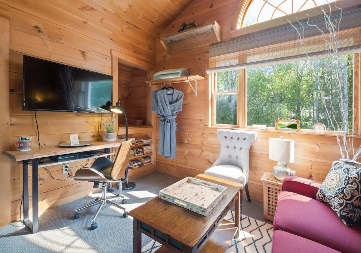 A warm and inviting sitting area features wood-paneled walls and large windows allowing natural light. A comfortable red sofa with patterned cushions faces a small table, while a desk with a chair provides a workspace. A flat-screen TV is mounted on the wall for entertainment.