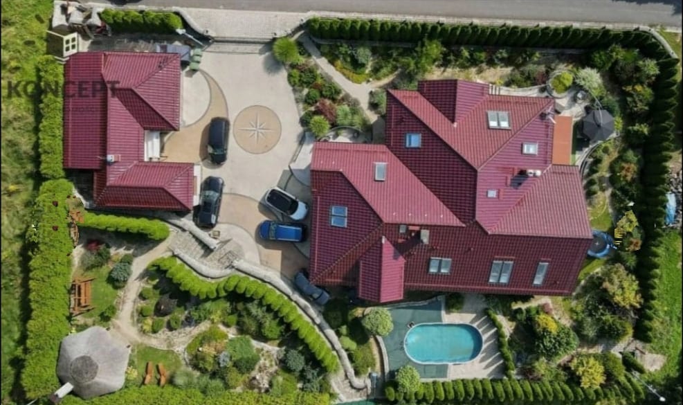 An aerial view captures a sprawling property featuring multiple buildings with red-tiled roofs. A well-maintained garden surrounds the structures, and a pool is visible amidst landscaped greenery. Driveways accommodate several vehicles, creating an organized arrangement within the space.