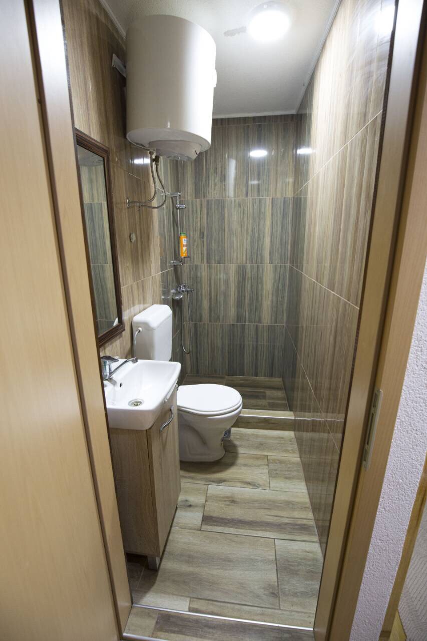 A compact bathroom is depicted, featuring warm wooden paneling and tiled flooring. A modern toilet and a small sink with a mirror above are visible. Shower fixtures are attached to the wall. Ample lighting is provided by ceiling fixtures.
