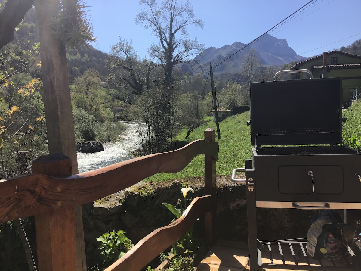 A barbecue grill is positioned on a wooden deck, overlooking a lush landscape with a flowing river and distant mountains. Trees provide a natural backdrop, and the clear blue sky enhances the outdoor space.