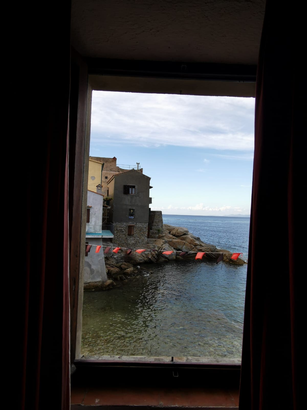 A view through an open window reveals a tranquil coastal scene, featuring colorful boats and rocky outcrops. The gentle waves reflect the sky, while charming buildings line the water's edge, showcasing varied architectural styles. Soft light enhances the serene atmosphere of the harbor.