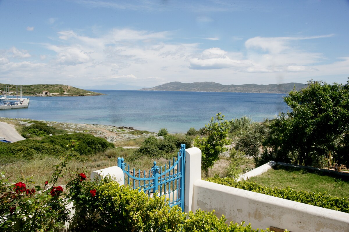 A view of the serene sea and distant hills is framed by lush greenery in the foreground. A blue gate leads into the property, surrounded by well-maintained gardens and blooming roses. The calming waters extend towards the horizon, providing a peaceful backdrop.