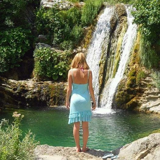 A person stands facing a gentle waterfall, surrounded by lush greenery. The clear water cascades over rocks, creating a small pool at the base. The vibrant colors of the natural landscape highlight the serene environment, creating a peaceful atmosphere in nature.